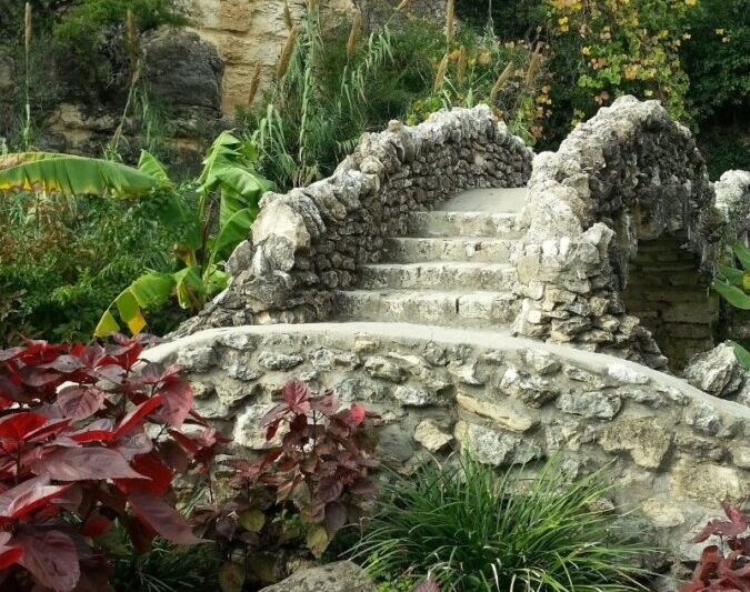 Stone bridge in lush garden with vibrant green plants and colorful flowers, Alamo Heights San Antonio Texas.