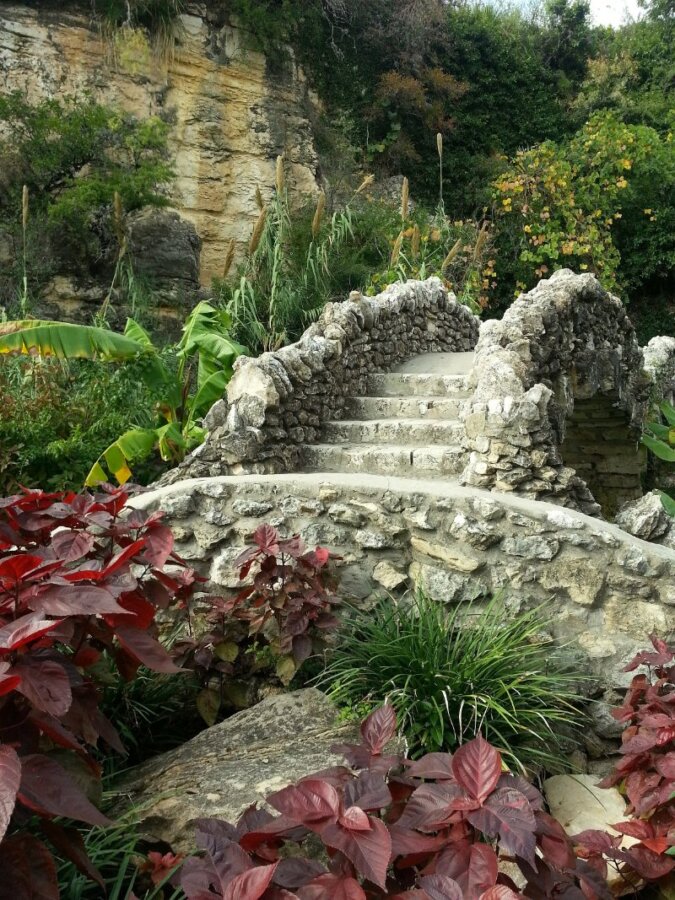 Stone bridge in lush garden with vibrant green plants and colorful flowers, Alamo Heights San Antonio Texas.