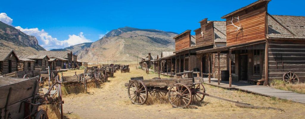Historic ghost town Cody, Wyoming, old wooden structures, abandoned wagons, mountainous backdrop.