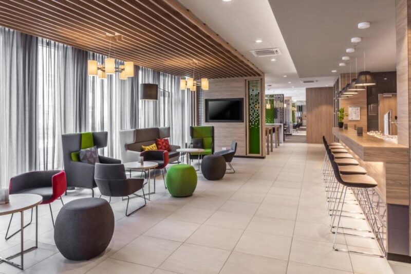 Here are three different alt text options for the given image 1. Spacious room with well-equipped bar, tables, chairs, and large TV at Holiday Inn Express Munich Ai City East 2. Cozy modern bar with alcoholic and non-alcoholic drinks, tables, chairs, and large TV at Holiday Inn Express Munich Ai City East 3. Lively room with well-stocked bar, tables, chairs, and large TV at Holiday Inn Express Munich Ai City East