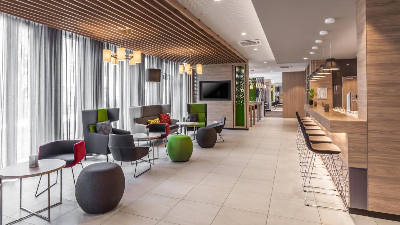 Here are three different alt text options for the given image 1. Spacious room with well-equipped bar, tables, chairs, and large TV at Holiday Inn Express Munich Ai City East 2. Cozy modern bar with alcoholic and non-alcoholic drinks, tables, chairs, and large TV at Holiday Inn Express Munich Ai City East 3. Lively room with well-stocked bar, tables, chairs, and large TV at Holiday Inn Express Munich Ai City East