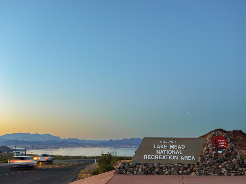 Dusk view of Lake Mead National Recreation Area entrance sign with passing cars and scenic backdrop.