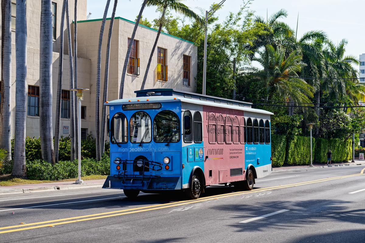 Miami Beach Trolley on a sunny day, featuring vibrant blue and pink colors, cruising past palm trees and art deco buildings.