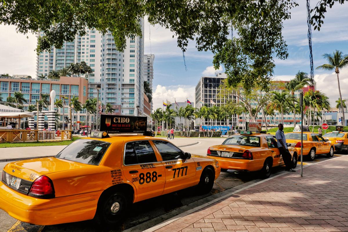 Yellow taxis lined up in downtown Miami with high-rise buildings and palm trees in the background.
