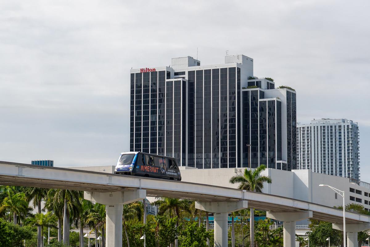 Miami Metromover train on elevated tracks with modern high-rise buildings and palm trees, including a prominent Hilton hotel.