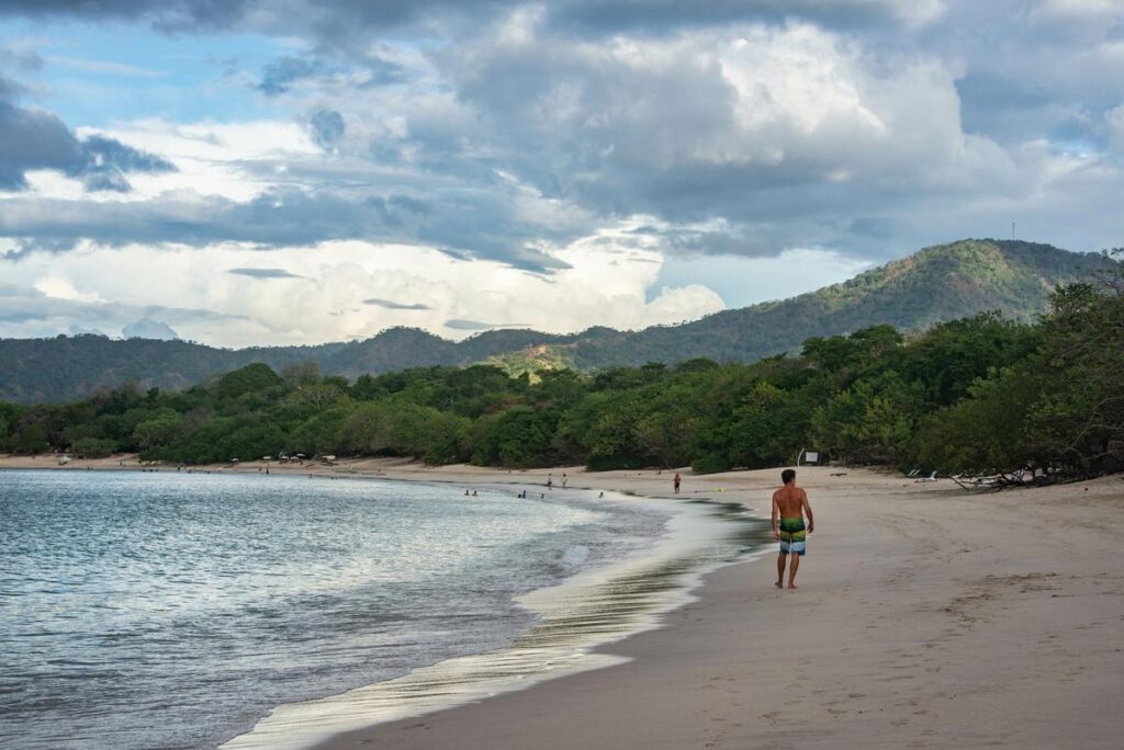 Tranquil Playa Brasilito beach scene with lone visitor, gentle waves, lush foliage, and majestic hills in Costa Rica.