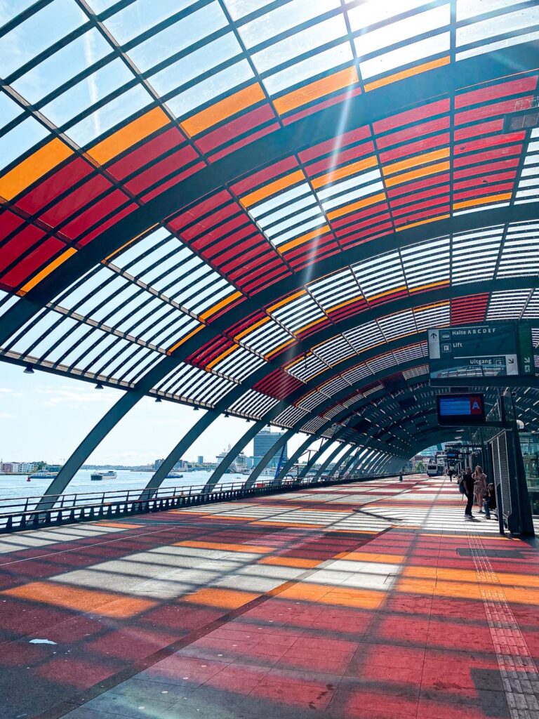Commuters at Amsterdam Centraal Station bus terminal with vibrant stained-glass roof.