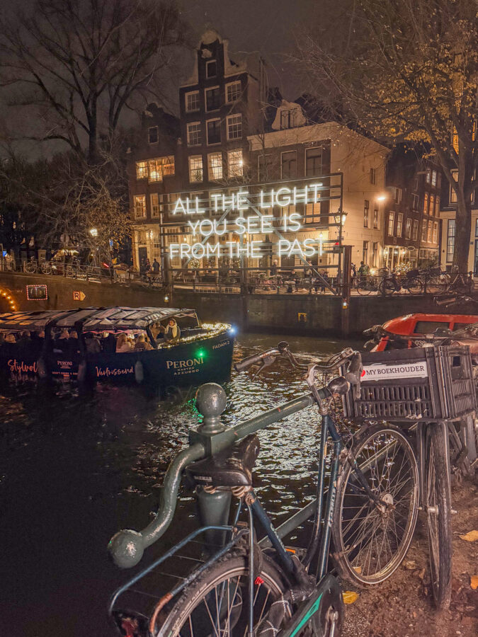 Amsterdam canal at night with neon sign, bicycle, boat, and Light Festival reflections.