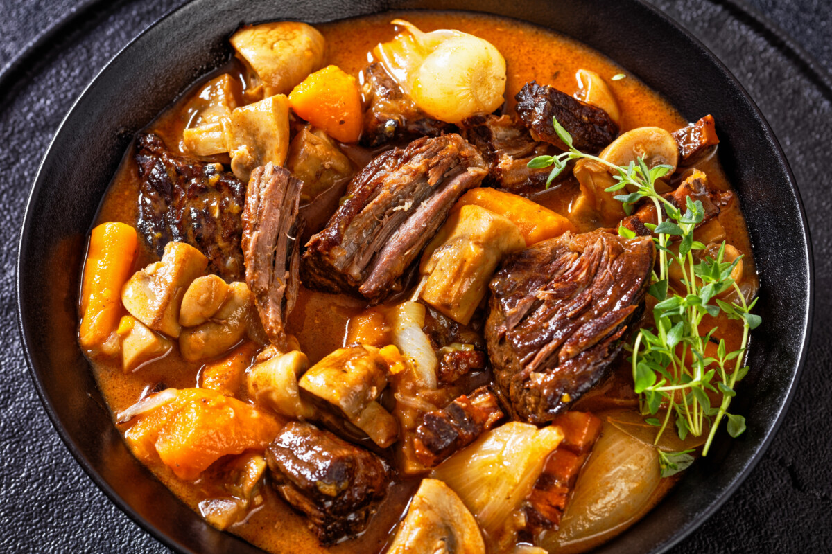 Close up of Beef Bourguignon, with beef, bacon, carrots, onion bulbs and mushrooms in rich red wine sauce in black bowl