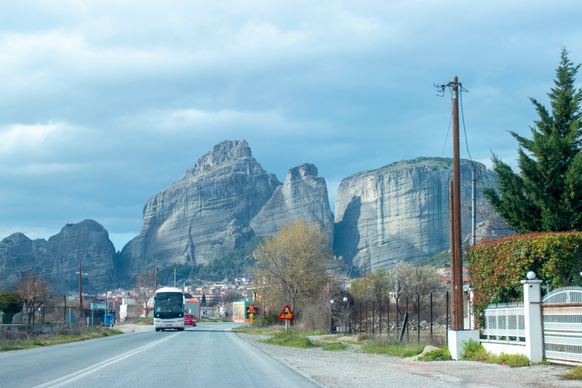 Bus on Kalampaka, Meteora, Greece rural road toward dramatic rock cliffs and small town beneath overcast sky.