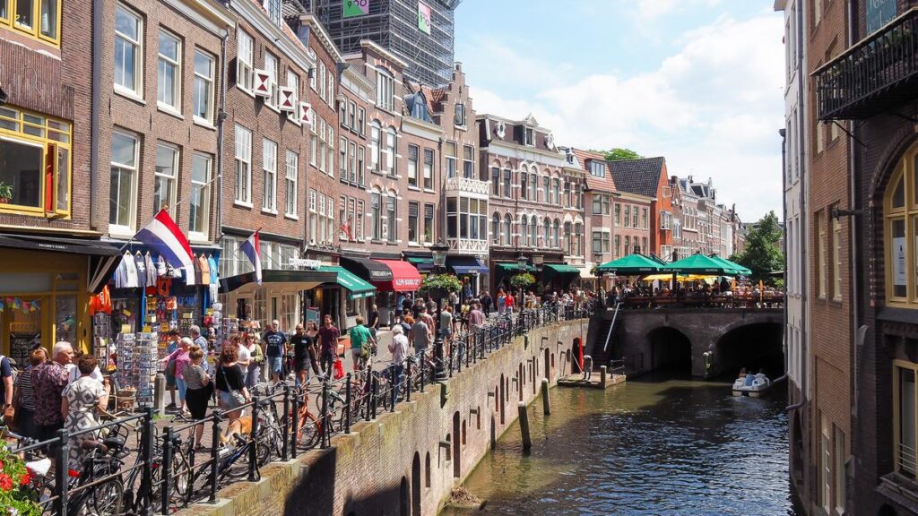 Bustling day at Oudegracht Canal, Utrecht on canal-side walkways, Dutch architecture, small businesses, and leisure boat.