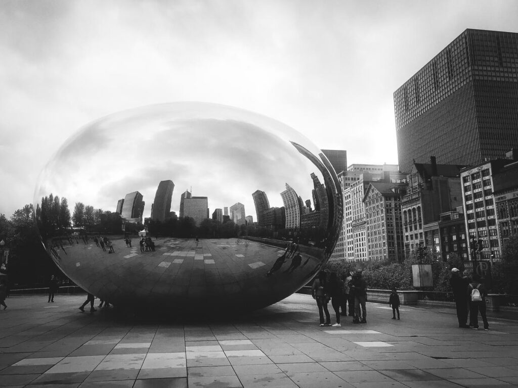 Black and white photograph of Cloud Gate sculpture in Millennium Park, Chicago, reflecting iconic city skyline and human interaction.