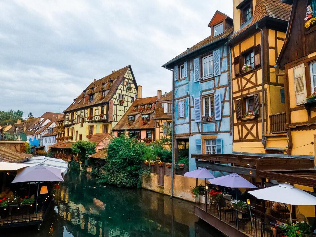 Charming La Petite Venise canal view in Colmar, France, with vibrant half-timbered houses and al fresco dining under pastel umbrellas, capturing Alsace's traditional architecture and romantic atmosphere.