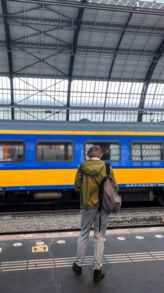 A man waits in front of a yellow and blue NS train at Amsterdam Centraal Station.