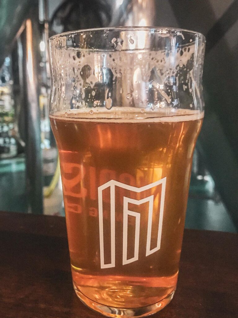 Half-full pint glass of amber beer with Modist Brewing Company logo in a Minneapolis brewery with soft focus background.
