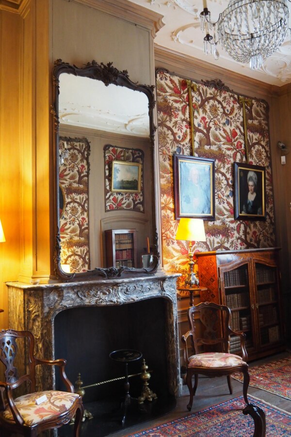Interior of a historical drawing room in Museum van Loon, featuring a carved fireplace, classic floral wallpaper, antique chairs, and oil paintings, under the soft glow of a chandelier.
