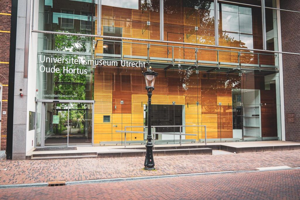 Exterior view of Universiteitsmuseum Utrecht Oude Hortus, showcasing modern glass facade with transparent entrance and bold lettering, framed by classic street lamp and cobblestone pavement.