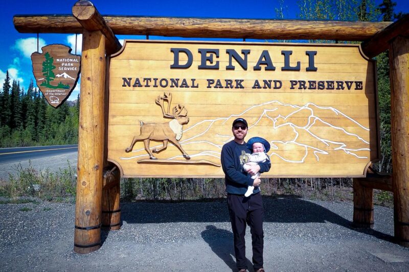 Kyle Kroeger and daughter at Denali National Park wooden entrance sign, showcasing park's iconic wildlife and vast landscapes, under clear blue skies.