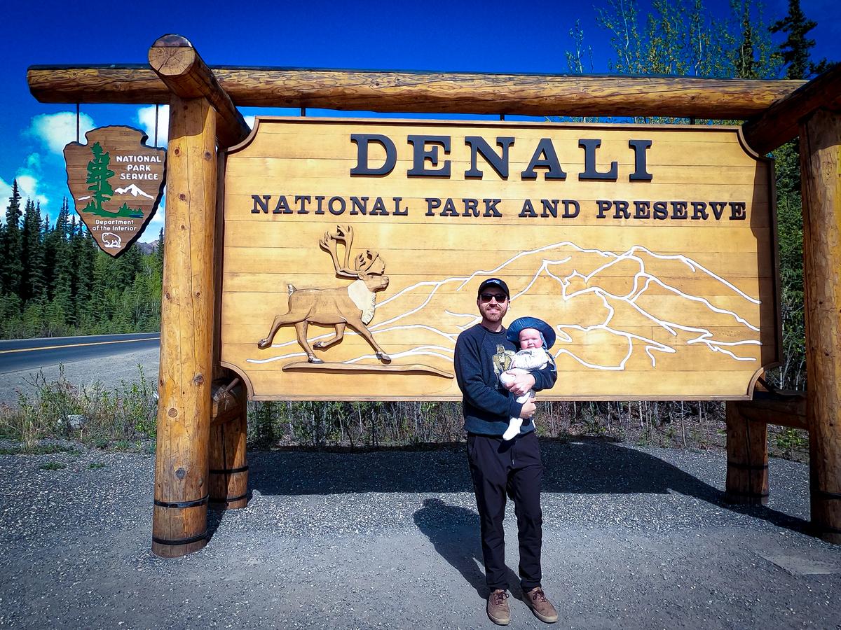 Kyle Kroeger and daughter at Denali National Park wooden entrance sign, showcasing park's iconic wildlife and vast landscapes, under clear blue skies.