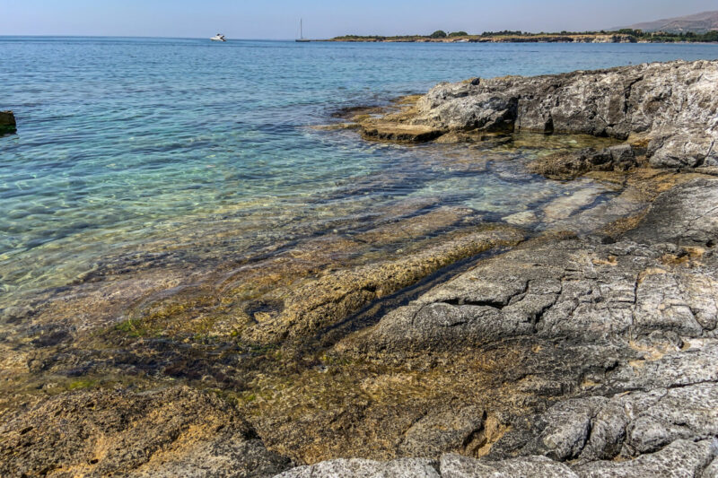 The alt text for this image should be "View of Fontane Bianche beach in Syracuse, Sicily with crystal-clear water lapping against a rocky shore surrounded by greenery."