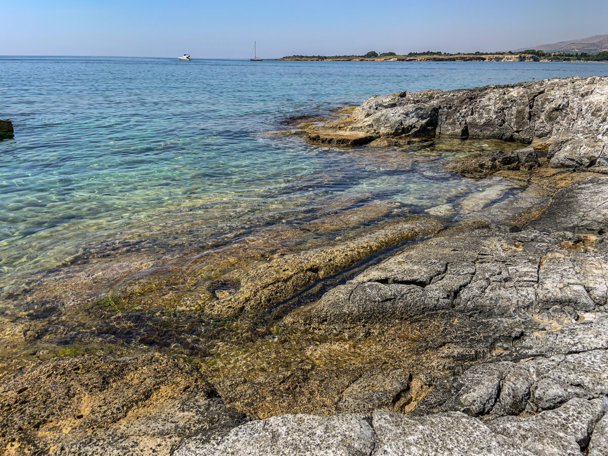 The alt text for this image should be "View of Fontane Bianche beach in Syracuse, Sicily with crystal-clear water lapping against a rocky shore surrounded by greenery."