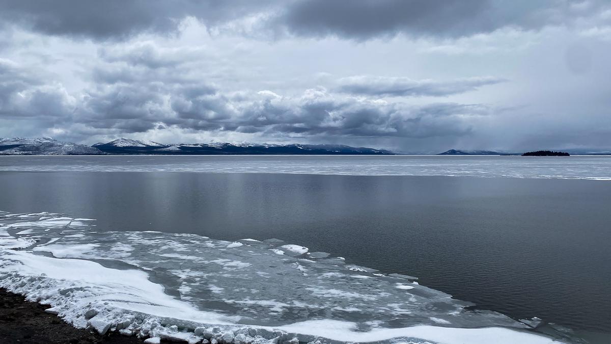 Partially frozen Yellowstone Lake under a cloudy sky with snow-covered mountains in the background, in early May.