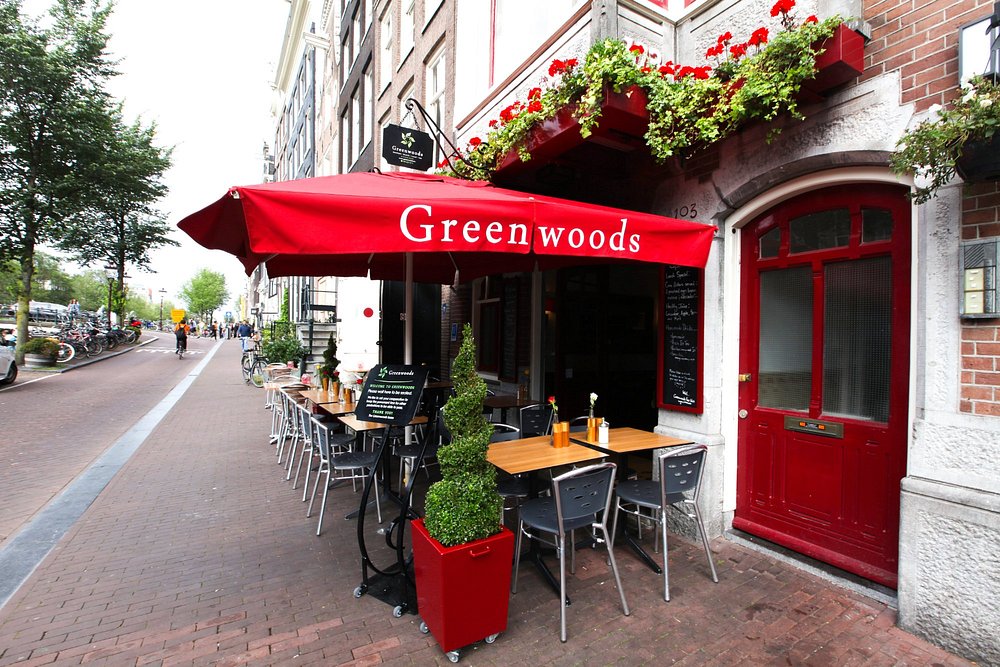 Greenwoods Singel Amsterdam sidewalk cafe with red canopy, wooden tables, metal chairs, brick facade, chalkboard, and planters.