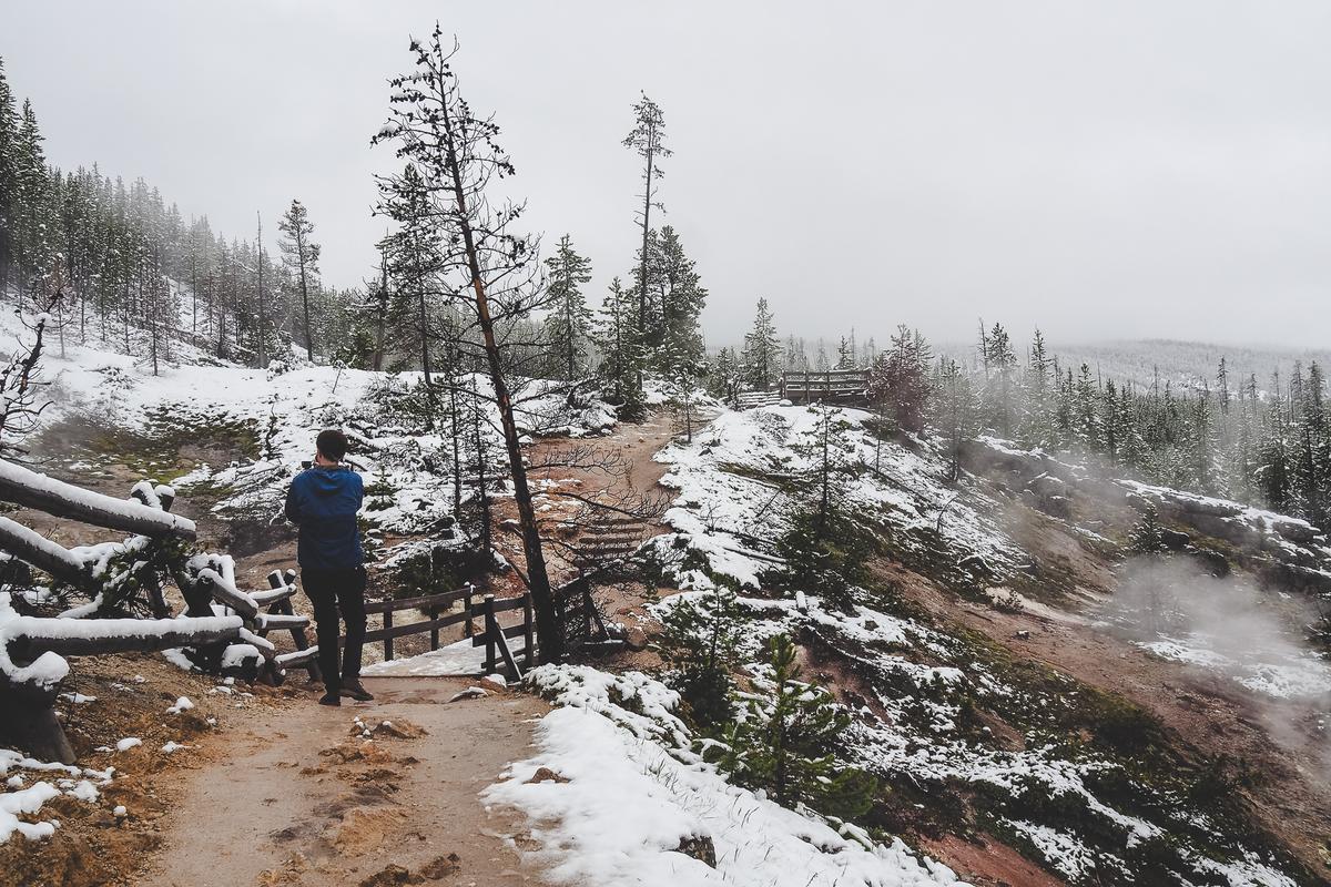 A hiker in a blue jacket observes steaming geothermal features along a snowy trail at Artist Paintpots, Yellowstone, surrounded by a wintry forest landscape.