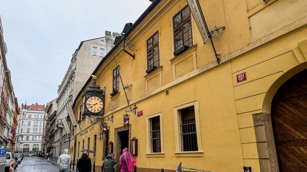 The historical U Fleků Beer Hall in Prague with its classic yellow exterior, ornate clock, and vintage signage, on a cobblestone street.