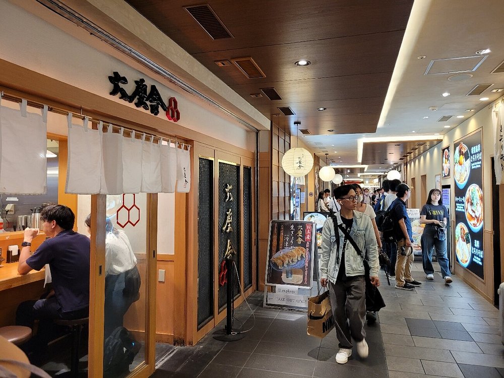Busy indoor Tokyo Ramen Street Japanese dining with counter seating, lanterns, illuminated menus, and warm wooden decor.