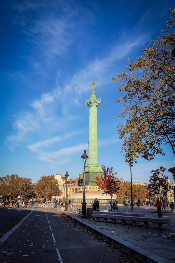 July Column at Place de la Bastille, Paris, in sunlight with autumn foliage and pedestrians.