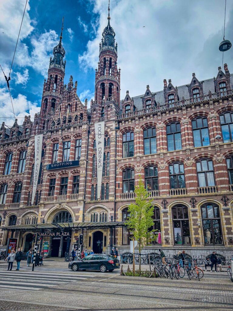 Magna Plaza, Amsterdam: Gothic Renaissance architecture, bustling street scene, cyclists, pedestrians, and clear blue sky.