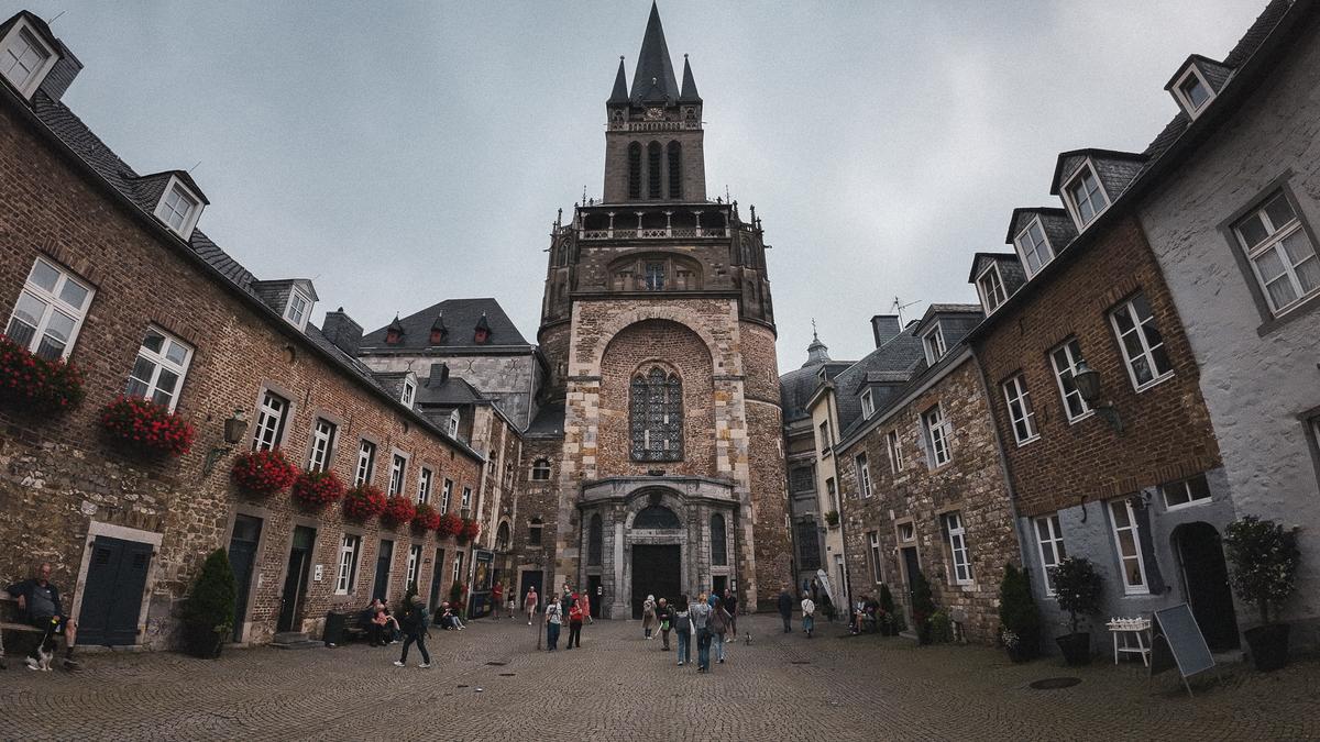 Atmospheric Aachen Cathedral entrance with historical buildings and people under a cloudy sky.