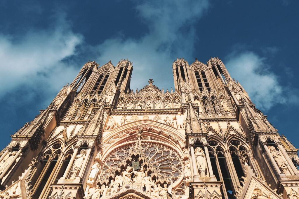 Ornate Gothic facade of Reims Cathedral with detailed sculptures and Rose window, set against a serene blue sky.