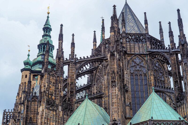 The Gothic spires and architectural details of St. Vitus Cathedral with contrasting turquoise roof under an overcast sky.