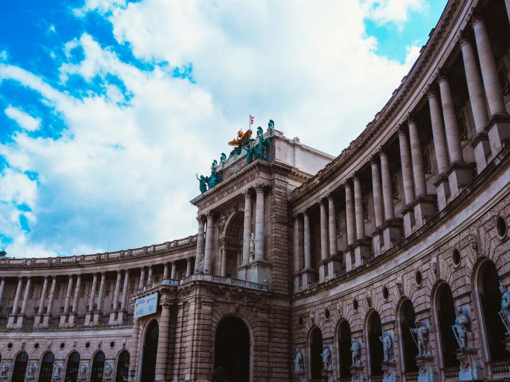 Exterior view of Hofburg Palace in Vienna with ornate sculptures and Corinthian columns under a blue sky with clouds.