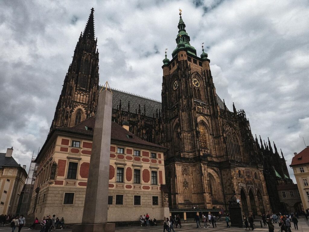 The intricate Gothic architecture of St. Vitus Cathedral in Prague with a modern obelisk in the foreground against a cloudy sky, capturing the essence of Czech history and architectural marvels.