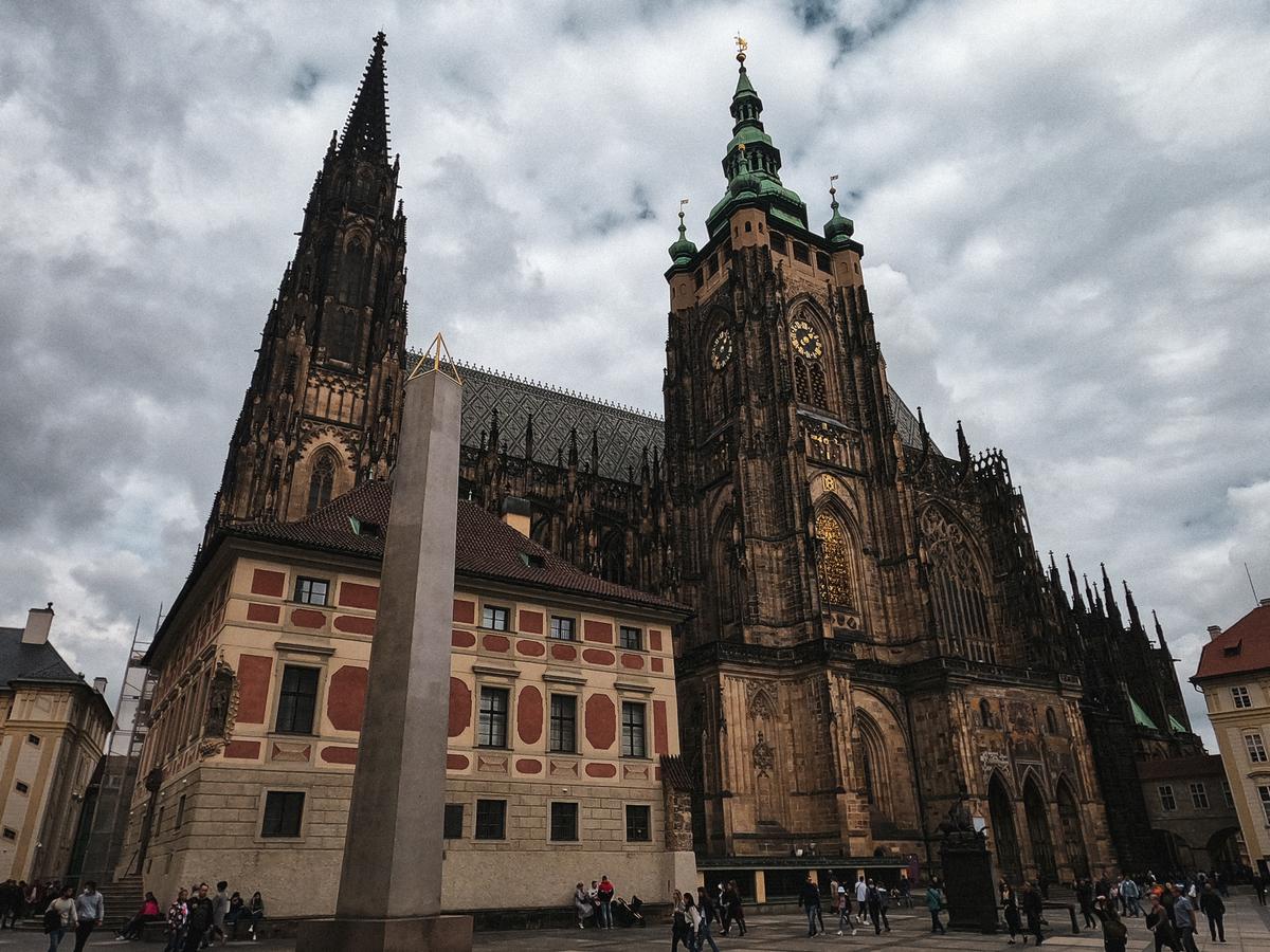 The intricate Gothic architecture of St. Vitus Cathedral in Prague with a modern obelisk in the foreground against a cloudy sky, capturing the essence of Czech history and architectural marvels.