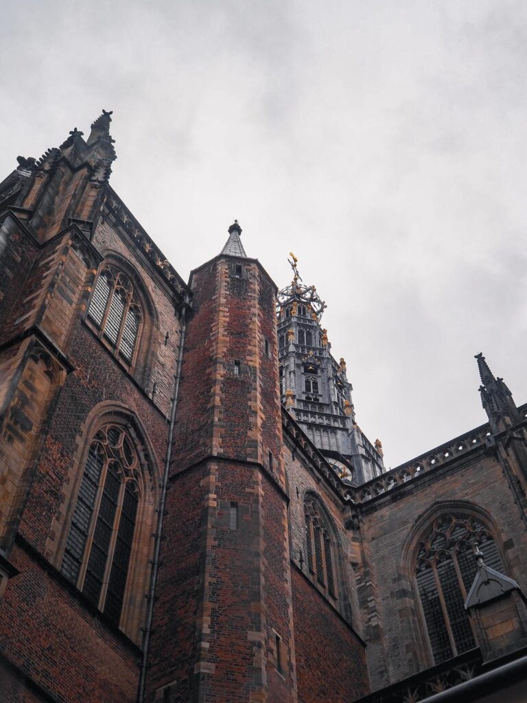 The soaring Gothic tower of St. Bavo Church in Haarlem, the Netherlands, with detailed brick patterns and a gold-topped weather vane, set against a moody gray sky.