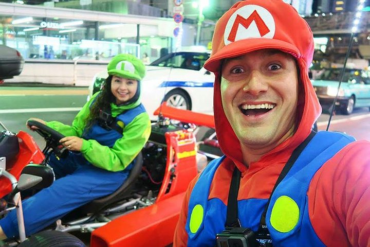 Go-Kart Tokyo! MariCAR Experience night city street selfie of Mario-inspired outfits beside a red go-kart amid traffic.