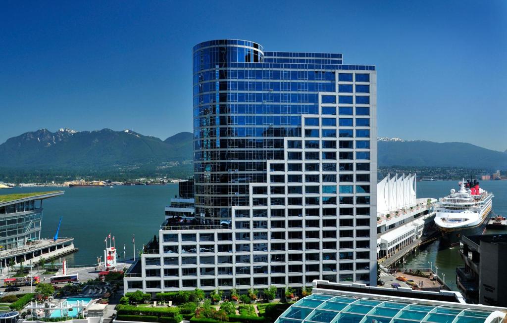 Fairmont Waterfront Hotel with glass facade overlooking harbor, cruise ship, sail canopies, terraces, mountains.