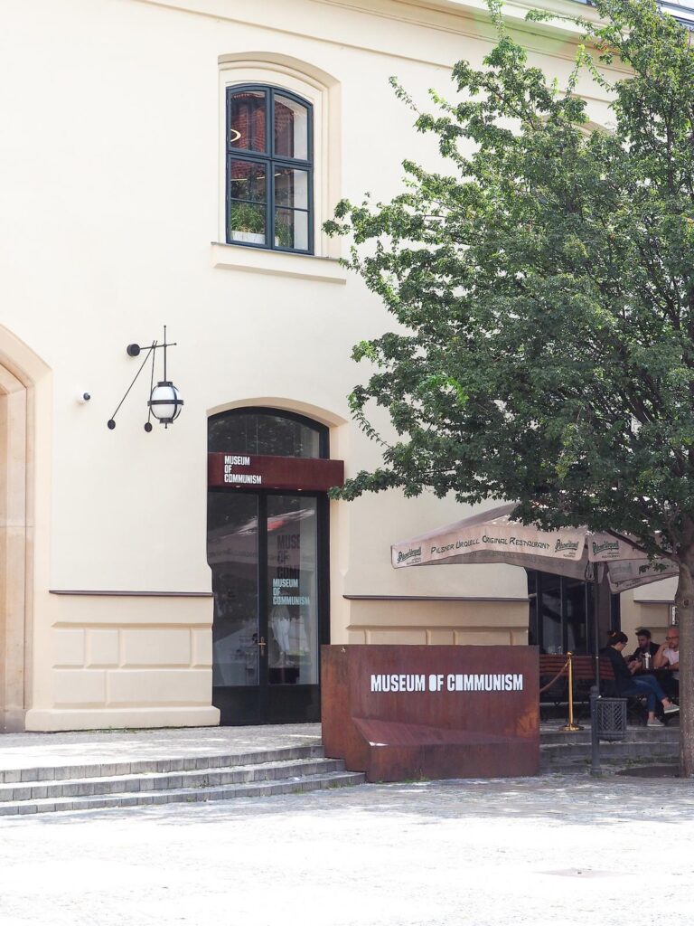 The front entrance of the Museum of Communism in Prague with a rust-colored sign, green foliage, and European architecture under a clear sky.