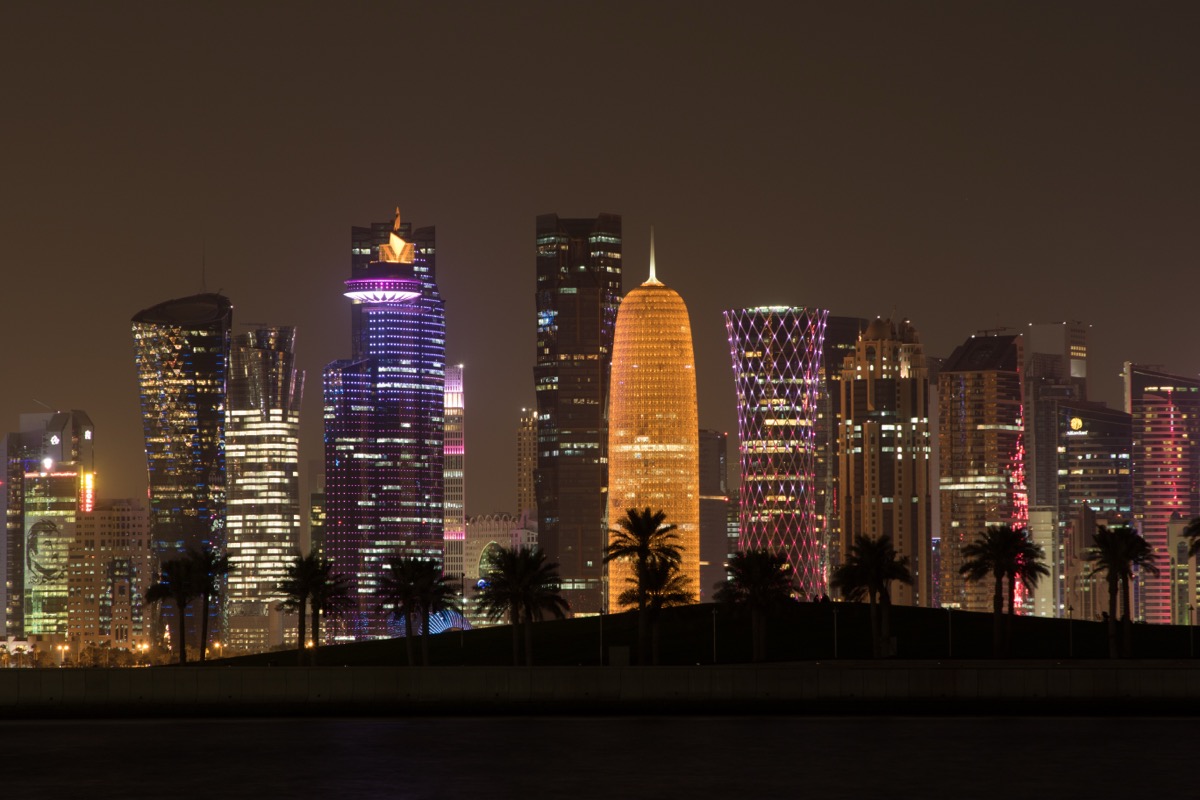 Doha, Qatar skyline at night with illuminated skyscrapers, amber lantern tower, and waterfront reflections.