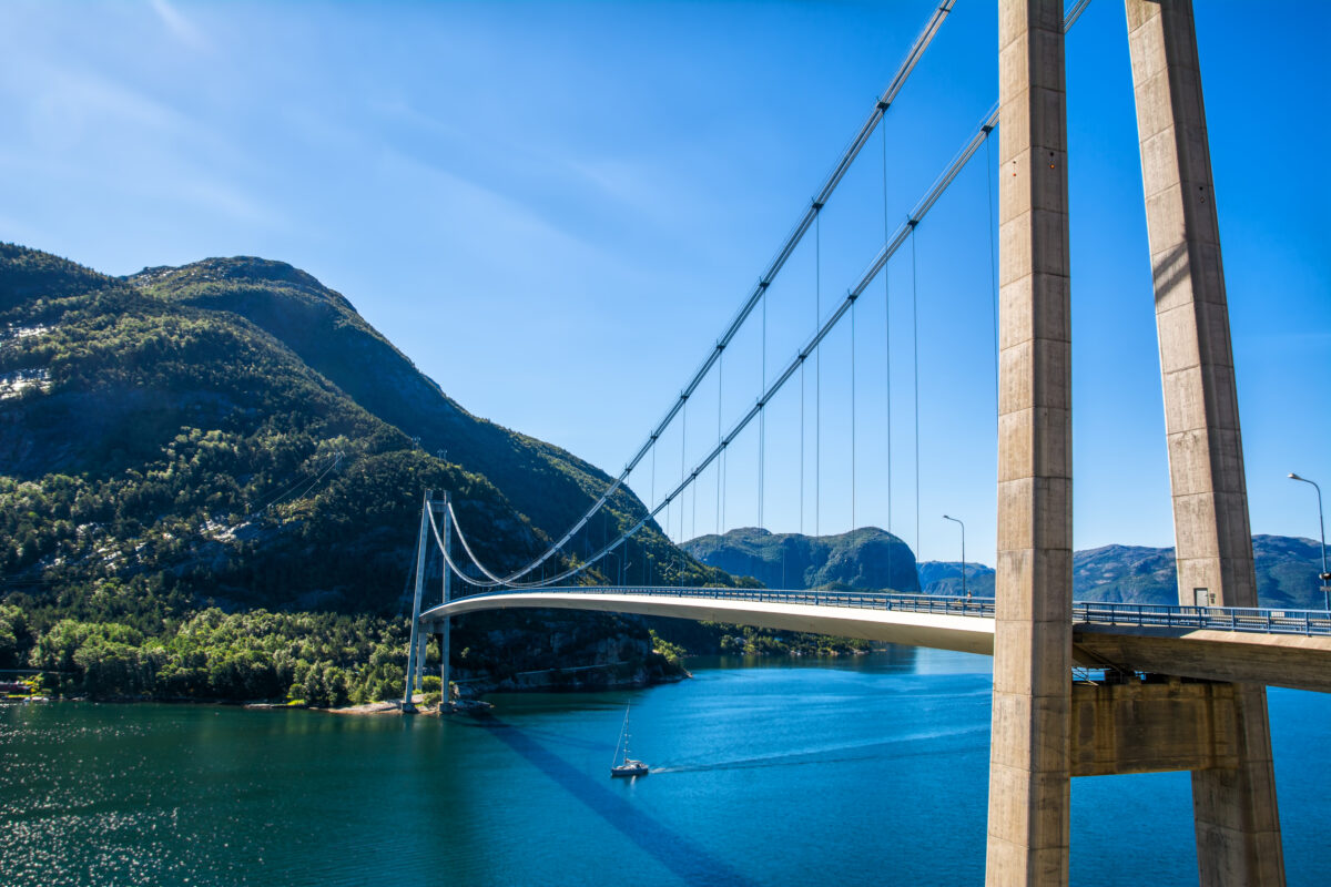 Norways Hardangerfjord suspension bridge spans a blue fjord, green mountains, and a sailing boat.
