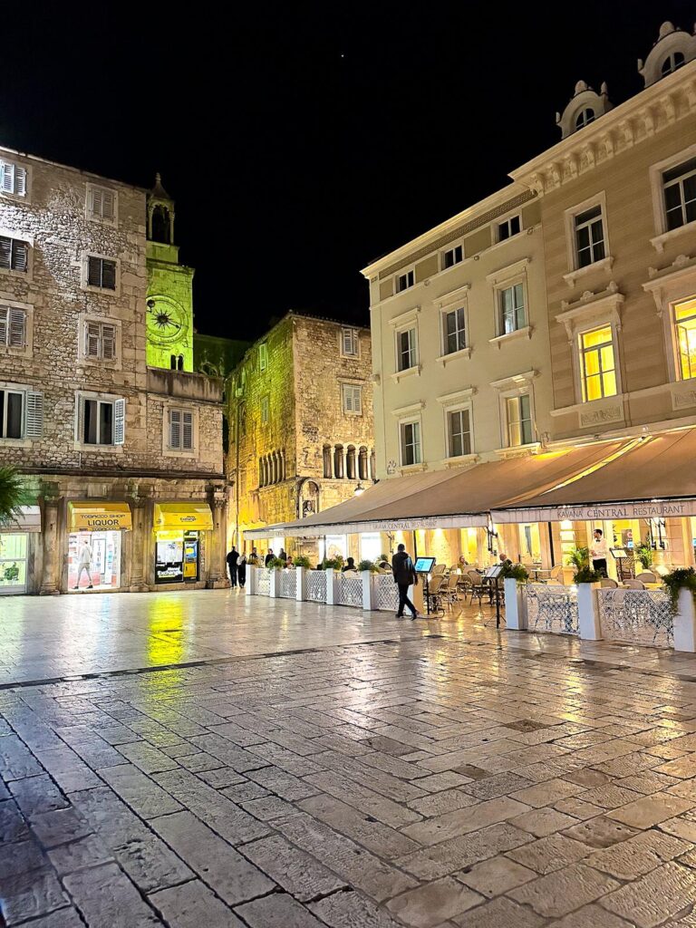 Night view of People's Square in Split, Croatia with illuminated historical buildings and outdoor dining.