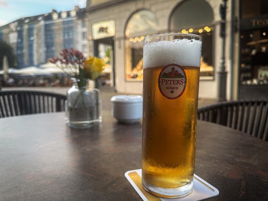 Chilled glass of Peters Kölsch beer on Aachen patio table with flower vase and blurred street scene.