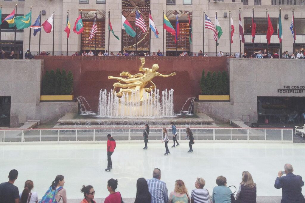 Skaters enjoying the Rockefeller Center Ice Skating Rink with the Prometheus statue and international flags in the background.