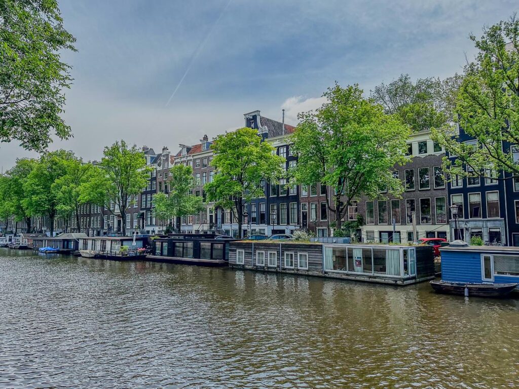 A vibrant capture of historic Dutch houses and houseboats along the Singel canal in Amsterdam, embraced by lush foliage and a tranquil watery expanse, under a soft spring sky.