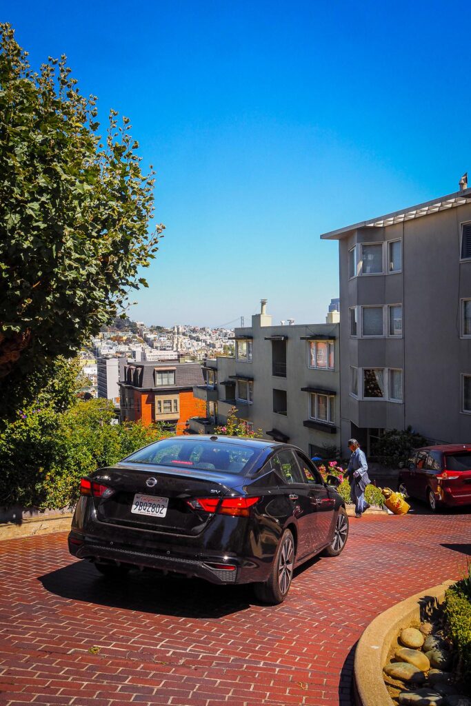 Scenic drive on Lombard Street, San Francisco with black sedan navigating iconic turns, colorful flowers, and lush greenery under clear blue sky.