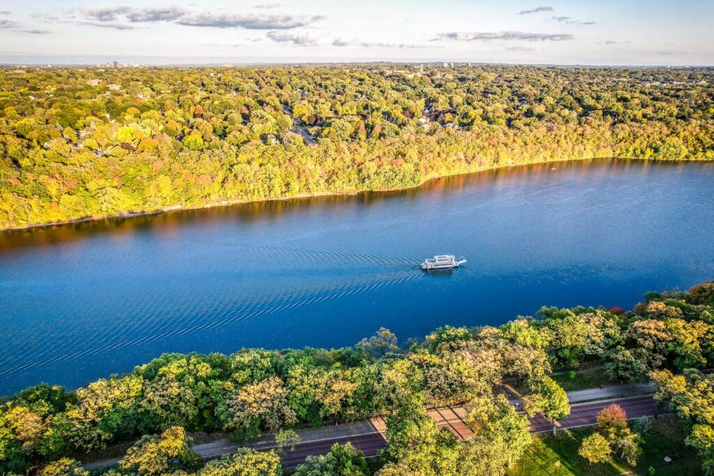 An aerial view of a leisure boat cruising along the Mississippi River, surrounded by autumn-hued trees and a clear blue sky, reflecting a tranquil and picturesque setting.